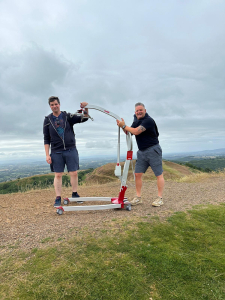 Two men stand atop a hill, proudly holding a mobile hoist against a clear sky.