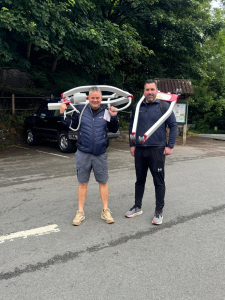 Two men stand by the roadside, each holding a mobile hoist part, engaged in conversation or waiting for someone.