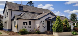 Exterior view of the Mill Race restaurant, a single-story building with white walls and dark roofing. Landscaped with shrubs in the foreground under a sunny sky.