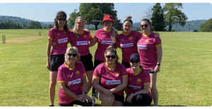 Group of cricket players in pink uniforms standing together on a sunny field, with scenic hills in the background. Goodrich Cricket Club.